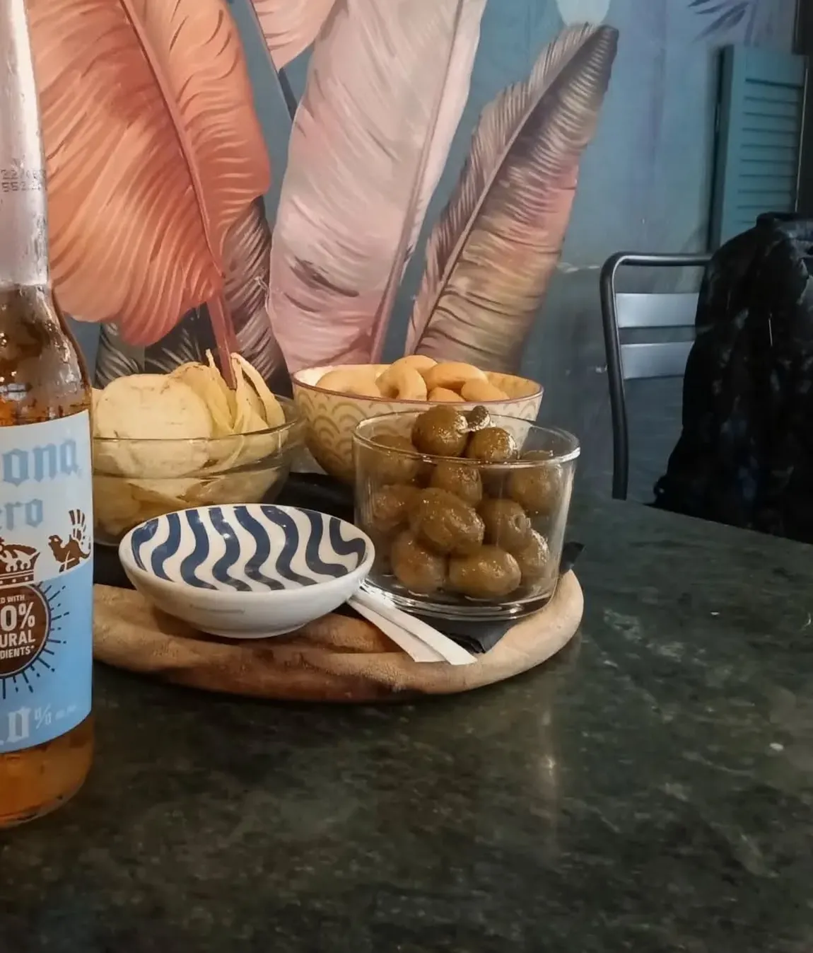 Tavolino con patatine, arachidi e olive davanti alla parete dipinta con grandi piume rosa e azzurre.