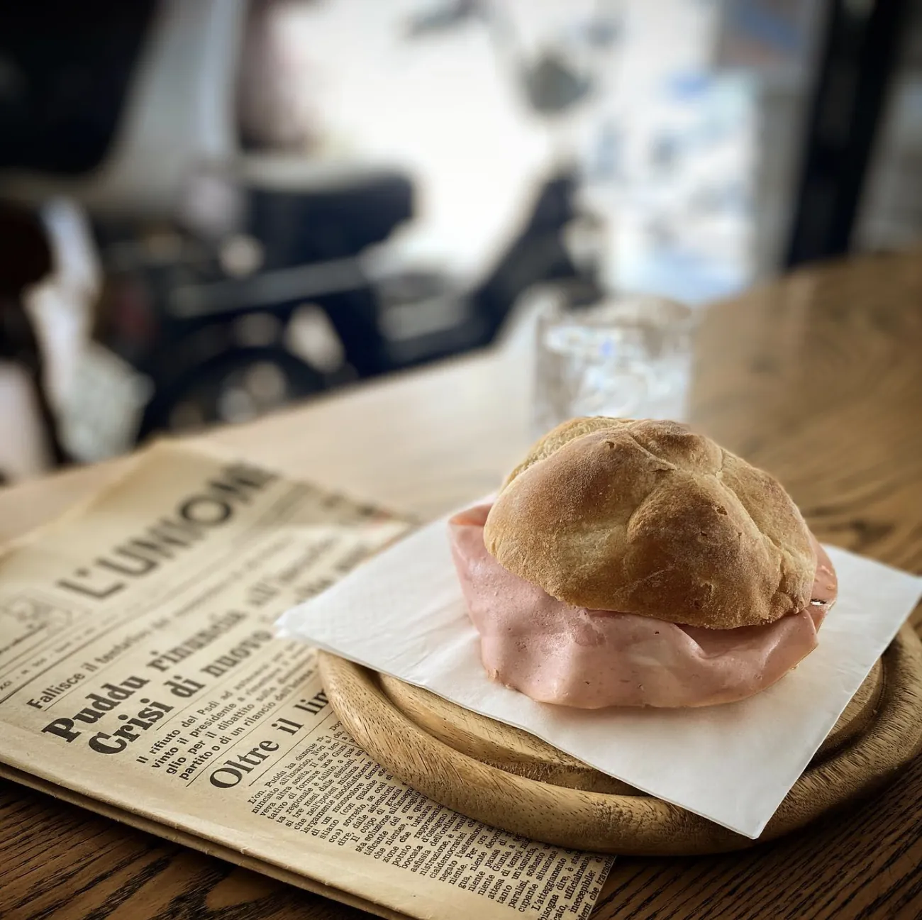 Panino con la mortadella su un tagliere di legno, appoggiato sopra una copia aperta del quotidiano L'Unione Sarda.