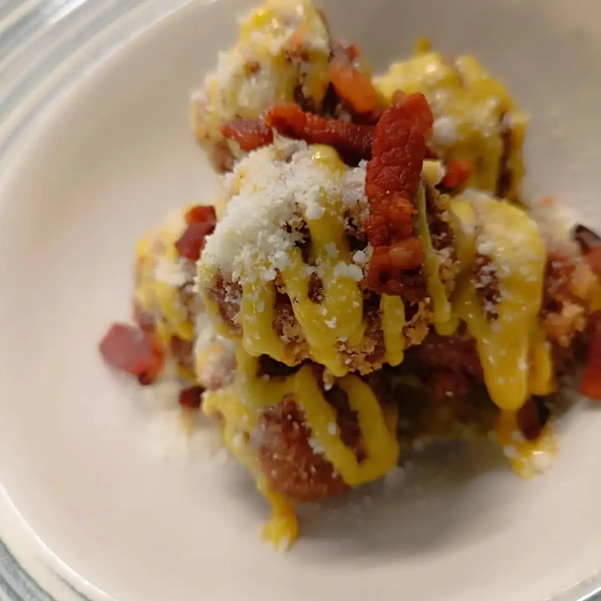 Piatto di polpette impanate con bacon croccante, salsa gialla e formaggio grattugiato.