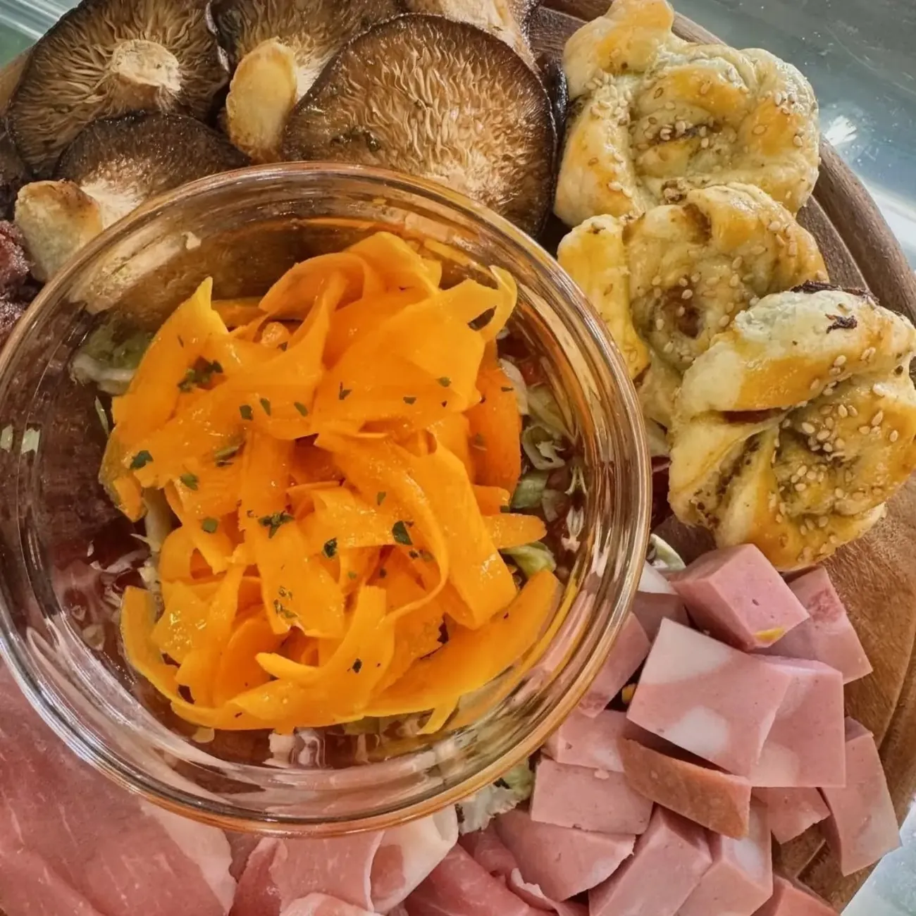 Tagliere di legno con salumi affettati, funghi grigliati, sfoglie al sesamo e una coppa di verdure.